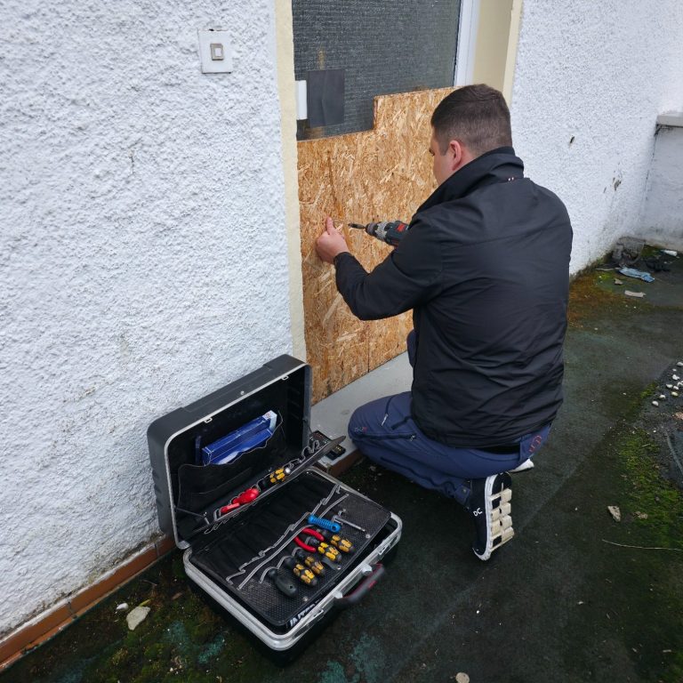Notverschalung nach einem Einbruch Mann befestigt ein Holzbrett vor einer Tür, Werkzeugkoffer neben ihm auf dem Boden.